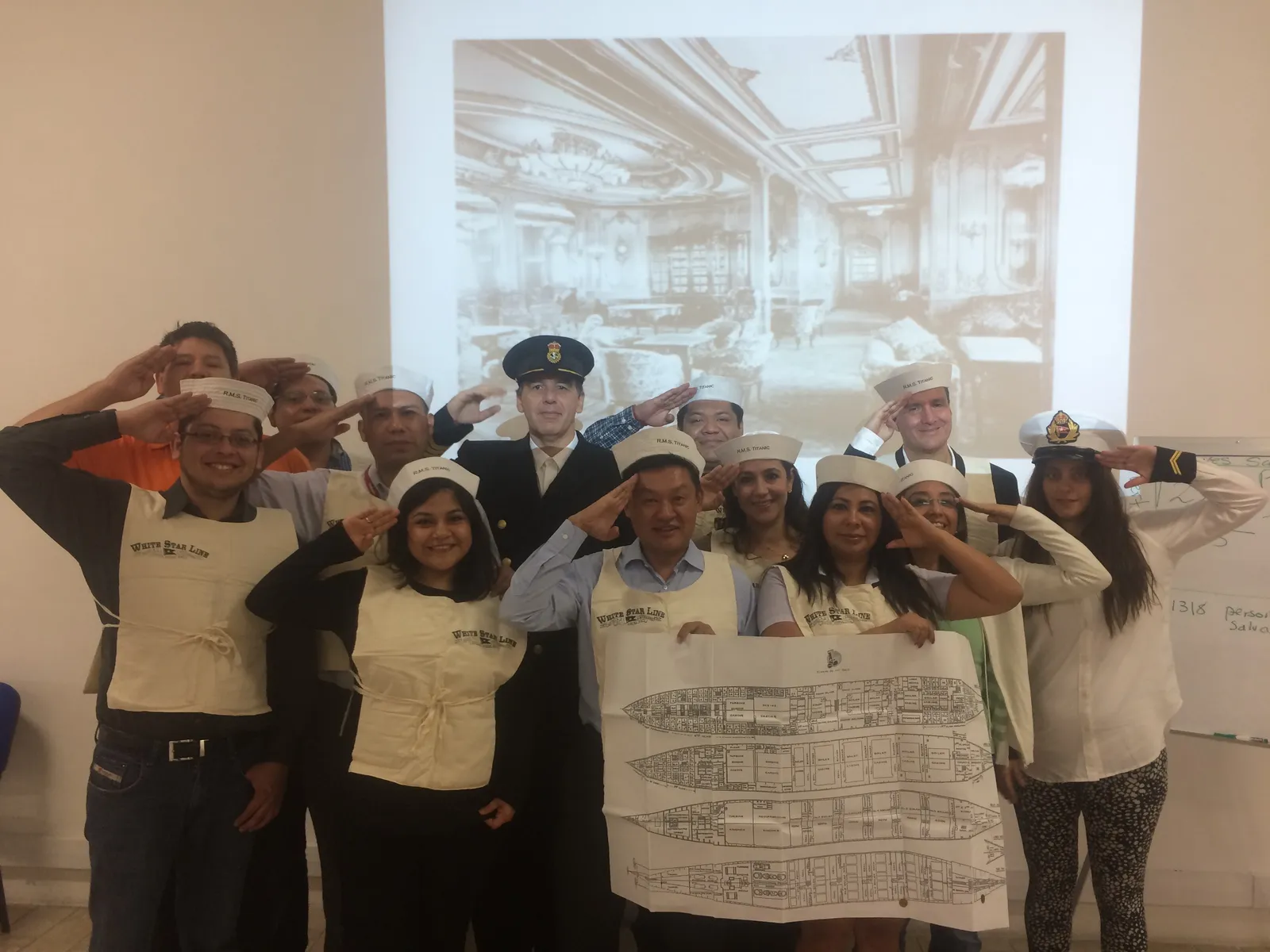 Leadership team saluting together after completing Save the Titanic experience, holding ship blueprints with Titanic interior projected behind them